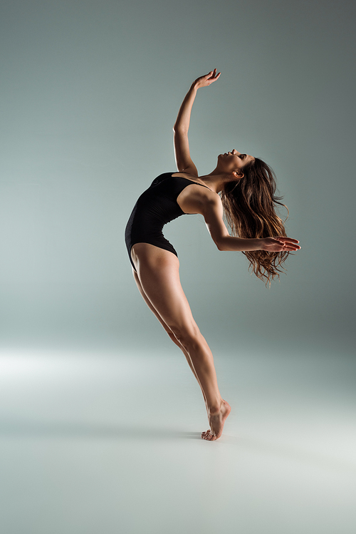 side view of attractive dancer in black bodysuit dancing contemporary on grey background