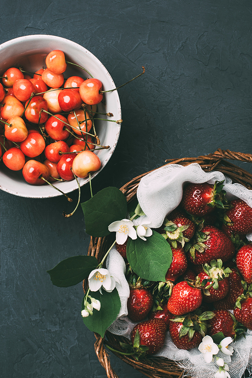 top view of sweet cherries in bowl and ripe strawberries with jasmine flowers in basket on black