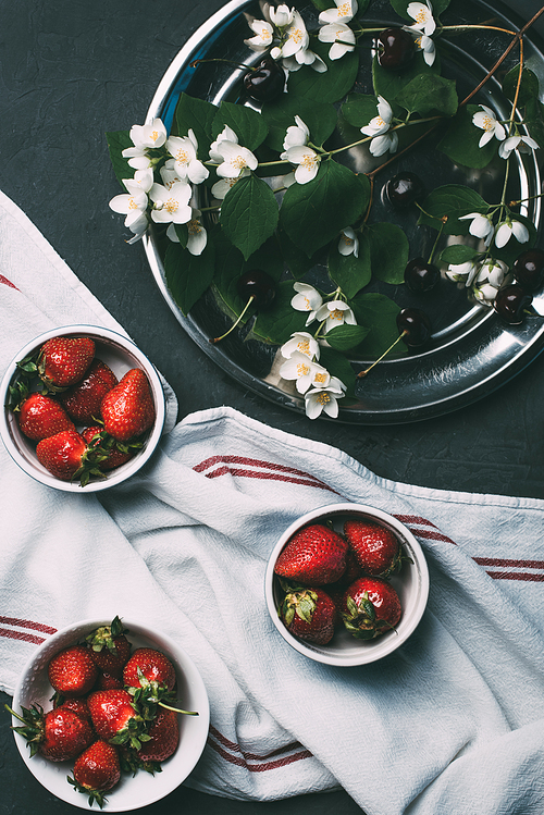 top view of ripe red strawberries in bowls, towel and jasmine flowers on black