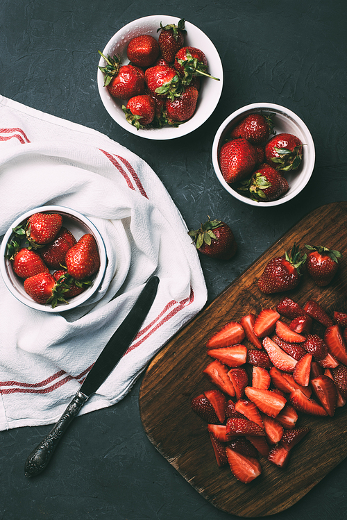 top view or ripe whole strawberries in bowls and sliced strawberries on wooden cutting board on black