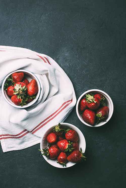 top view of ripe red strawberries in bowls and towel on black