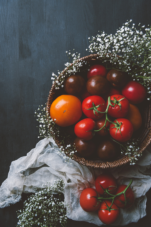 top view of fresh red tomatoes in wicker bowl with white flowers on wooden background