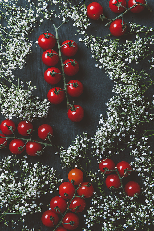 top view of red cherry tomatoes and white flowers on black wooden background
