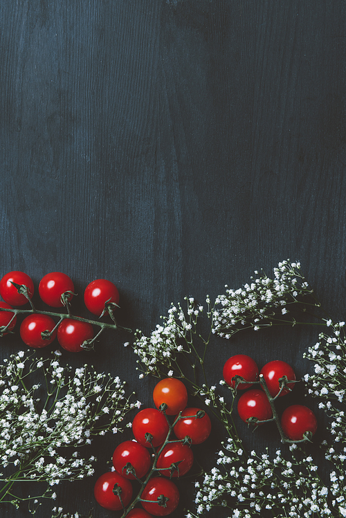 top view of ripe cherry tomatoes and white flowers on black wooden table with copy space
