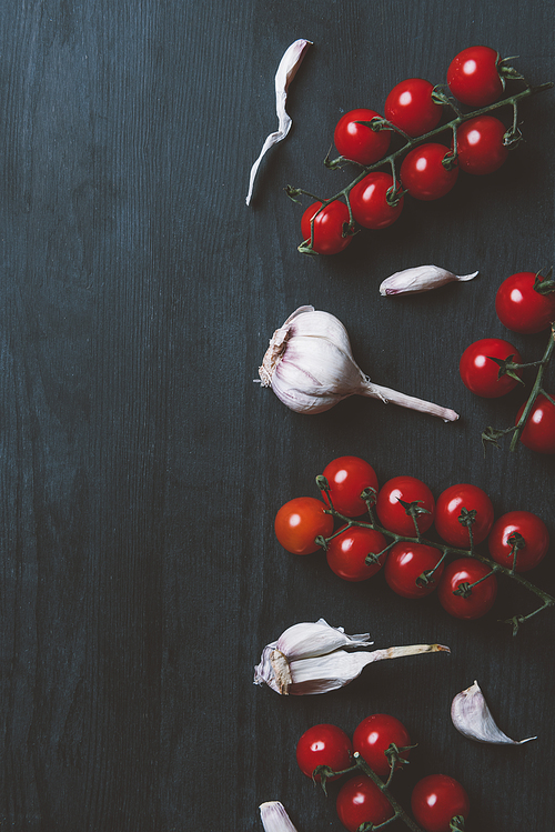 top view of red cherry tomatoes and garlic on black wooden background