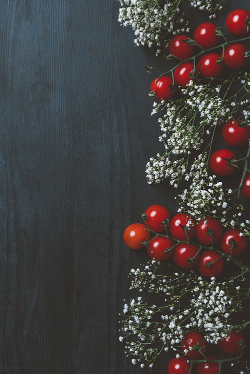 top view of red cherry tomatoes and white flowers on black wooden background