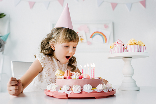 little birthday girl in cone blowing out candles from cake on table