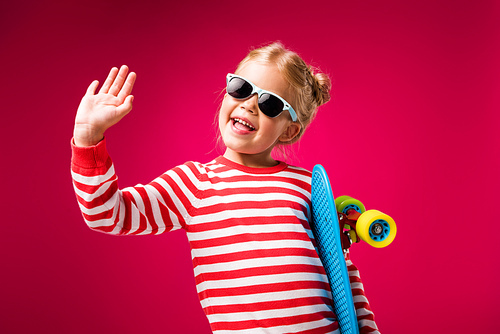 excited stylish kid in sunglasses holding penny board and waving isolated on red