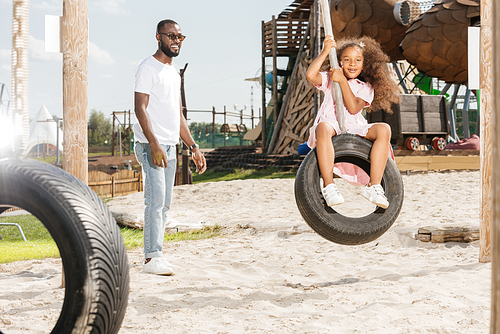 african american father standing near smiling daughter on tire swing at amusement park