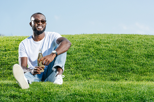 happy handsome african american man sitting with soda in glass bottle on green hill