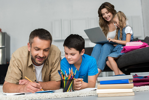 selective focus of father and son doing homework together while mother and daughter using laptop on sofa at home