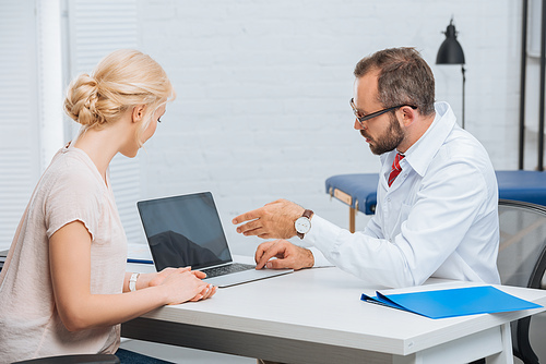 physiotherapist in white coat having conversation with female patient at workplace with laptop in hospital