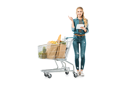 attractive young woman with digital tablet doing idea gesture and standing near shopping trolley with products isolated on white