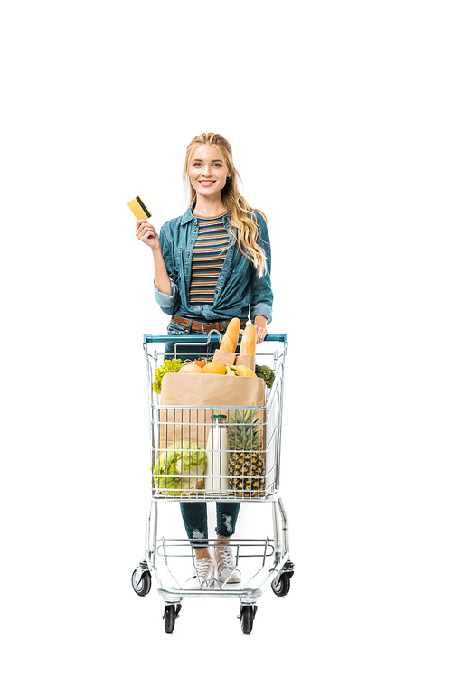 cheerful woman showing credit card and carrying shopping trolley with products isolated on white