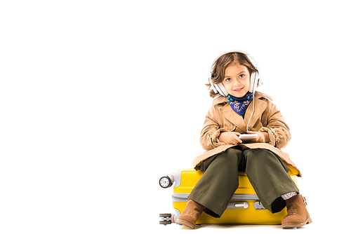 beautiful little child in trench coat listening music with headphones and sitting on yellow suitcase isolated on white