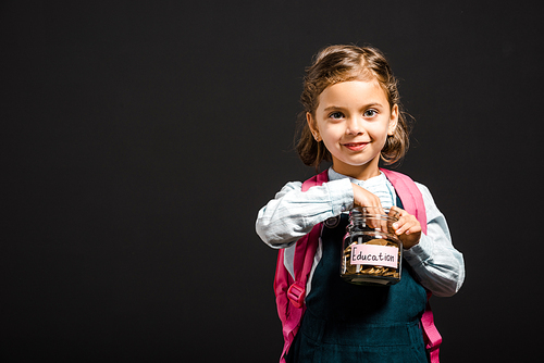 schoolgirl with backpack holding glass jar with savings for education isolated on black