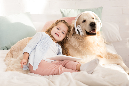 adorable kid holding tablet and leaning on cute golden retriever with headphones lying on bed in children room