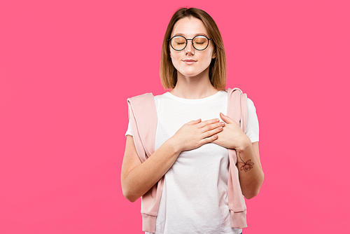 dreamy girl in eyeglasses holding hands on chest isolated on pink
