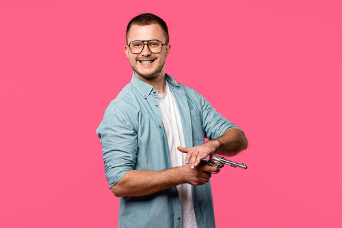 young man in eyeglasses holding revolver and smiling at camera isolated on pink