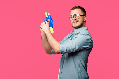 young man in eyeglasses holding toy gun and smiling at camera isolated on pink