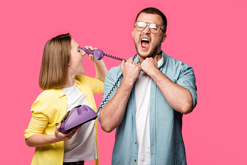 young woman talking by rotary phone and looking at man with wire around neck screaming isolated on pink