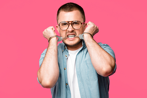 aggressive young man in eyeglasses biting handcuffs and  isolated on pink