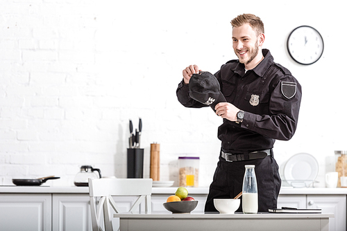 handsome police officer with cap in hands smiling at kitchen