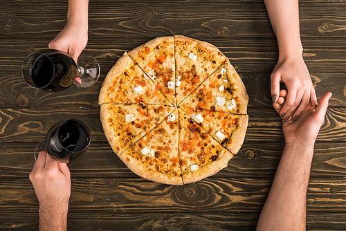 cropped view of couple holding hands and glasses of red wine while eating pizza on wooden table