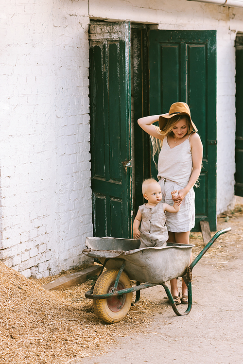 woman holding sons hand while he standing in wheelbarrow at countryside
