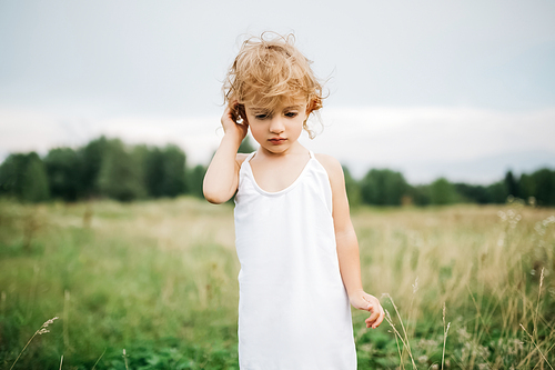 adorable child with curly hair standing in field and looking down