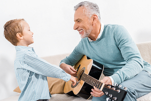 laughing middle aged man teaching grandson playing on acoustic guitar at home