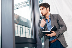 thoughtful businessman standing by window and holding notebook in office