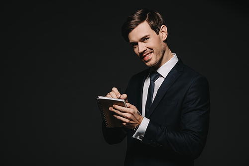 cheerful businessman writing in notebook with pen isolated on black