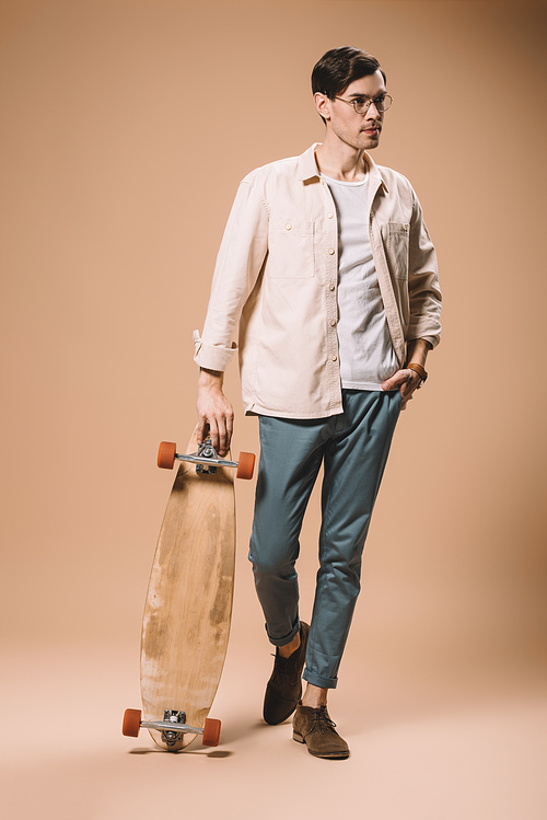 man in glasses holding skateboard while standing with hand in pocket isolated on beige
