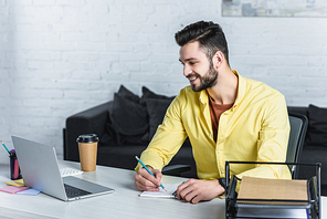 Smiling businessman using laptop and writing in notebook