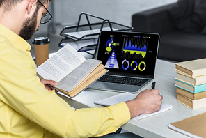 bearded man studying with book near laptop with charts and graphs on screen in modern office