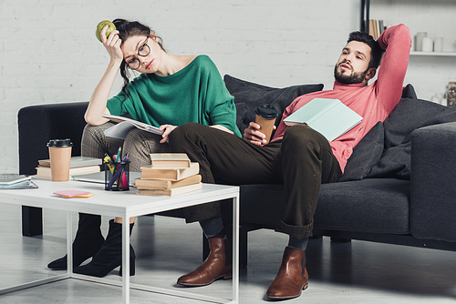 woman in glasses holding apple in hand and sitting near tired man with paper cup