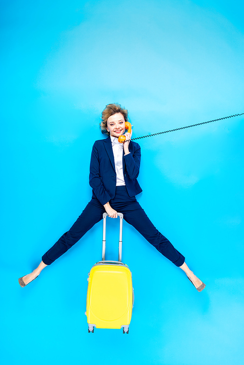Charming woman in suit with yellow valise talking on telephone on blue background