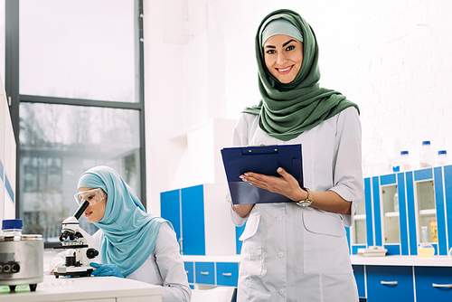 female muslim scientists using microscope and clipboard during experiment in chemical laboratory