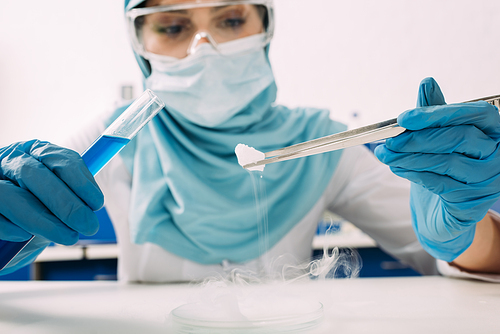 cropped view of female muslim scientist holding test tube with tweezers and experimenting with dry ice in laboratory