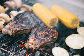 selective focus of juicy tasty steaks grilling on bbq grid with mushrooms, corn and sliced eggplant