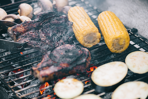 selective focus of tweezers and juicy tasty steaks grilling on bbq grid with mushrooms, corn and sliced eggplant