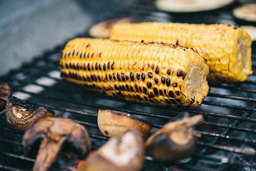 selective focus of yellow corn with crust and mushrooms grilling on barbecue grid