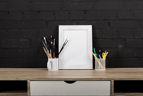 close up view of empty photo frame, paintbrushes and office supplies on table