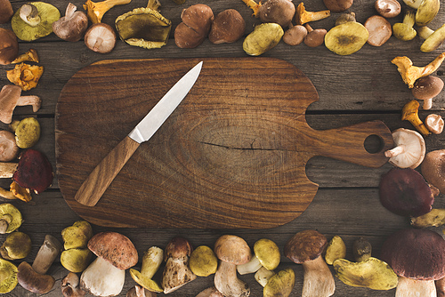 Top view of wooden cutting board with knife and mushrooms around on a table