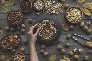 top view of human hand and various nuts in bowls with dried autumn leaves on table