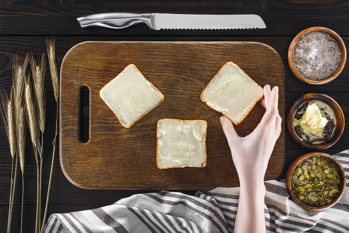 top view of human hand holding fresh toast with butter from wooden cutting board