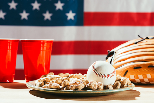 close-up view of baseball ball on plate with peanuts, red plastic cups and baseball glove on table with us flag behind
