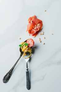 elevated view of spoon and fork wrapped by pasta with mint leaves, pine nuts, jamon and cherry tomatoes covered by grated parmesan on marble table