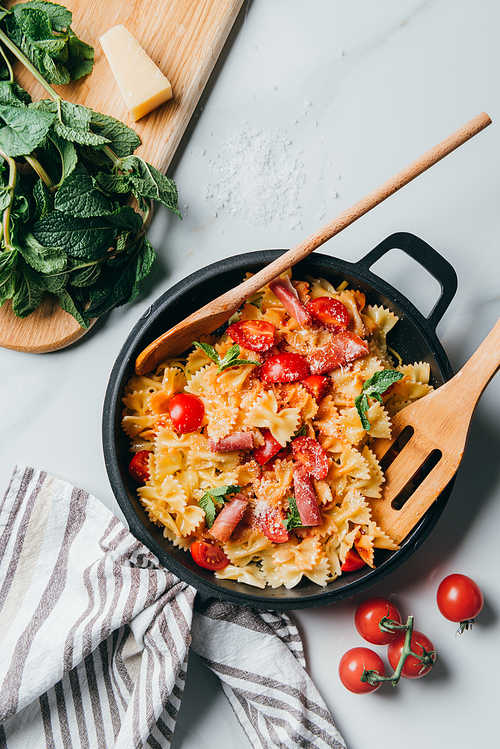 elevated view of pasta with jamon, cherry tomatoes, mint leaves covered by grated parmesan in pan with wooden spatula and spoon on marble table